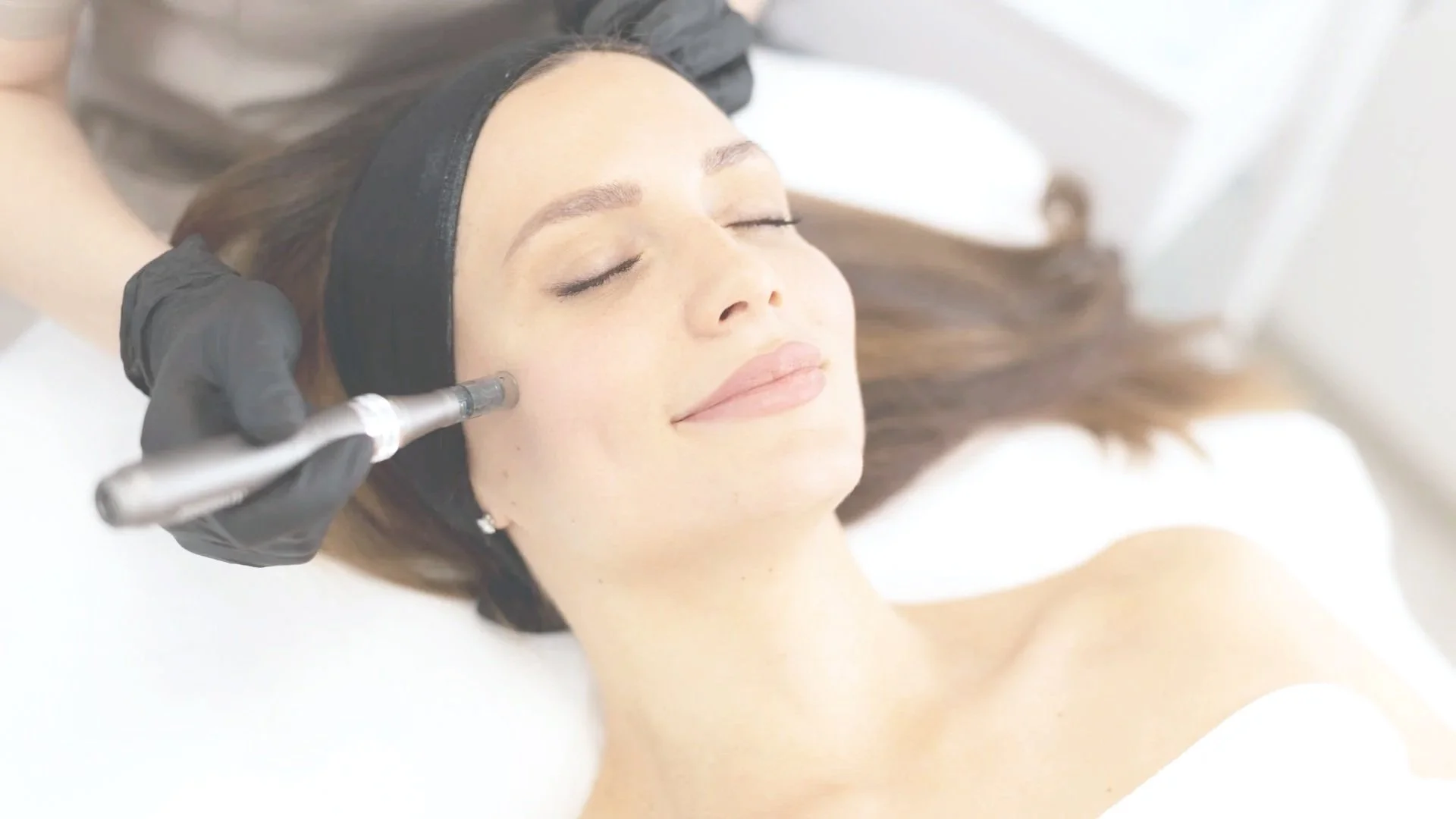A woman with closed eyes and a gentle smile receiving a facial treatment with a small handheld device on her face while lying down with a black headband, in a bright, clean setting.