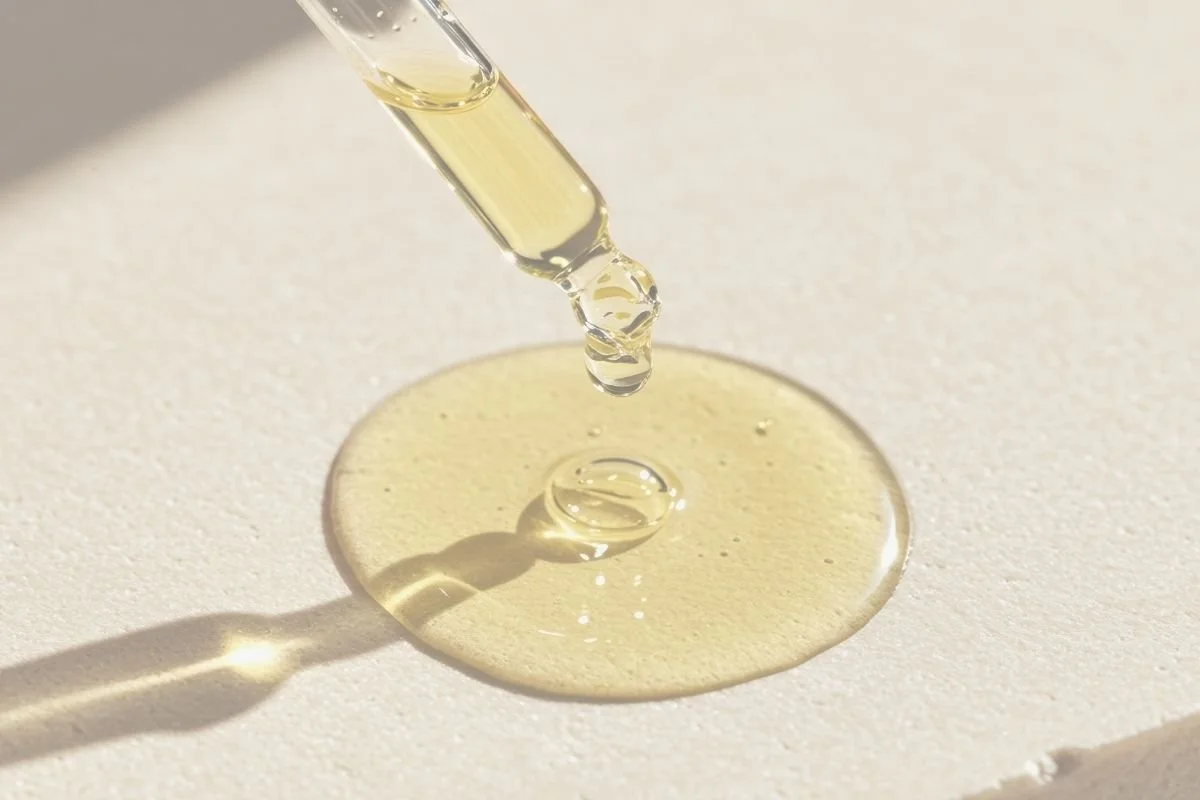 Close-up of a dropper dispensing clear oil onto a round drop of oil on a white surface.