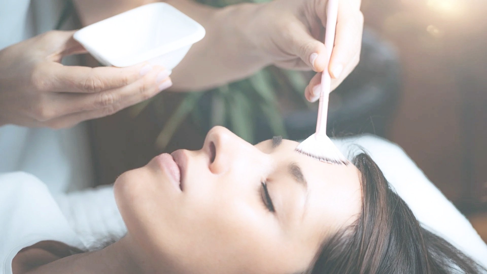 A woman lying down with her eyes closed while a person prepares to apply makeup to her face using a cotton swab.