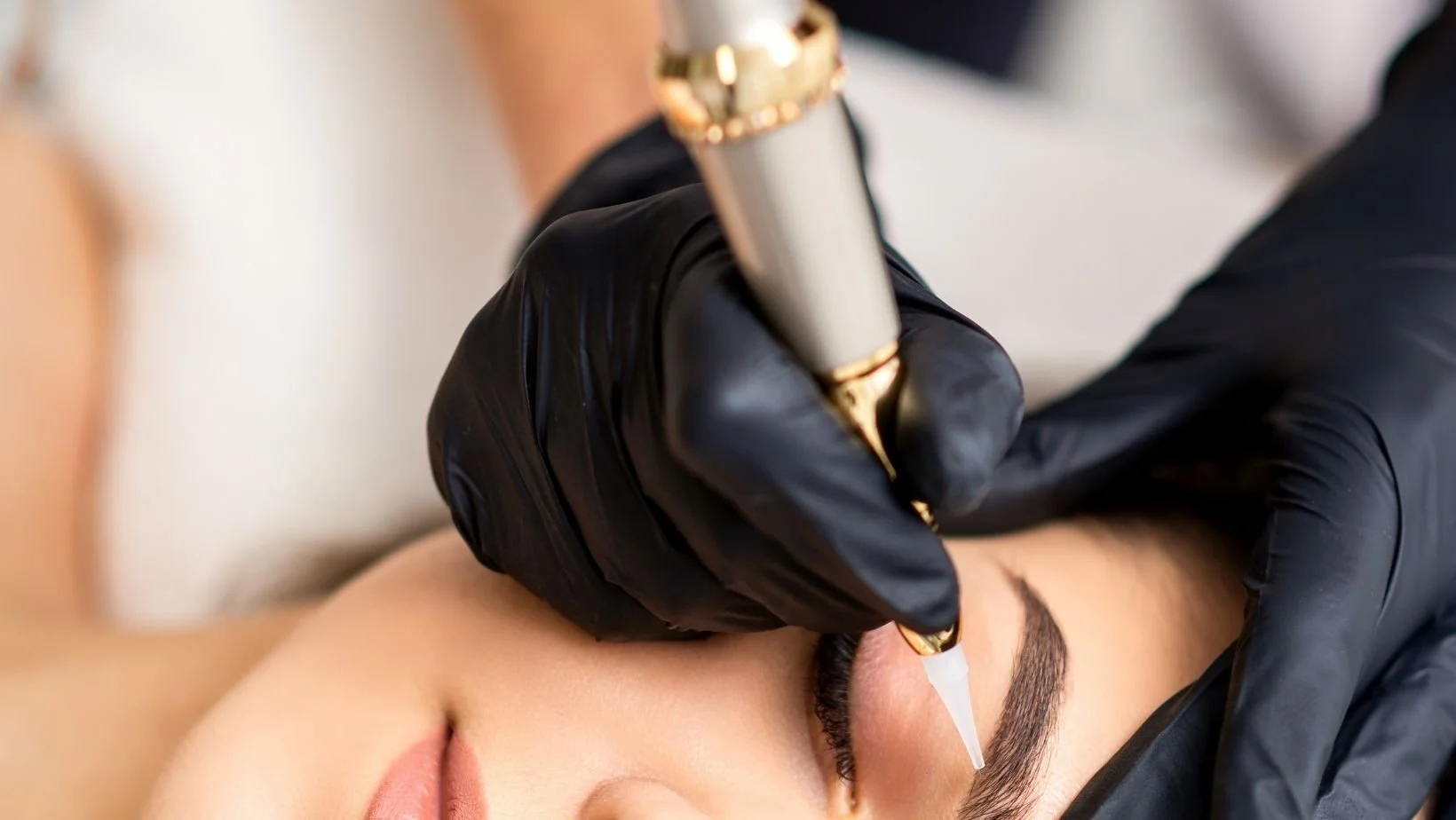 A person receiving a cosmetic eyebrow tattooing procedure, lying down with eyes closed, while a technician in black gloves uses a pen-like instrument to create permanent eyebrow makeup.