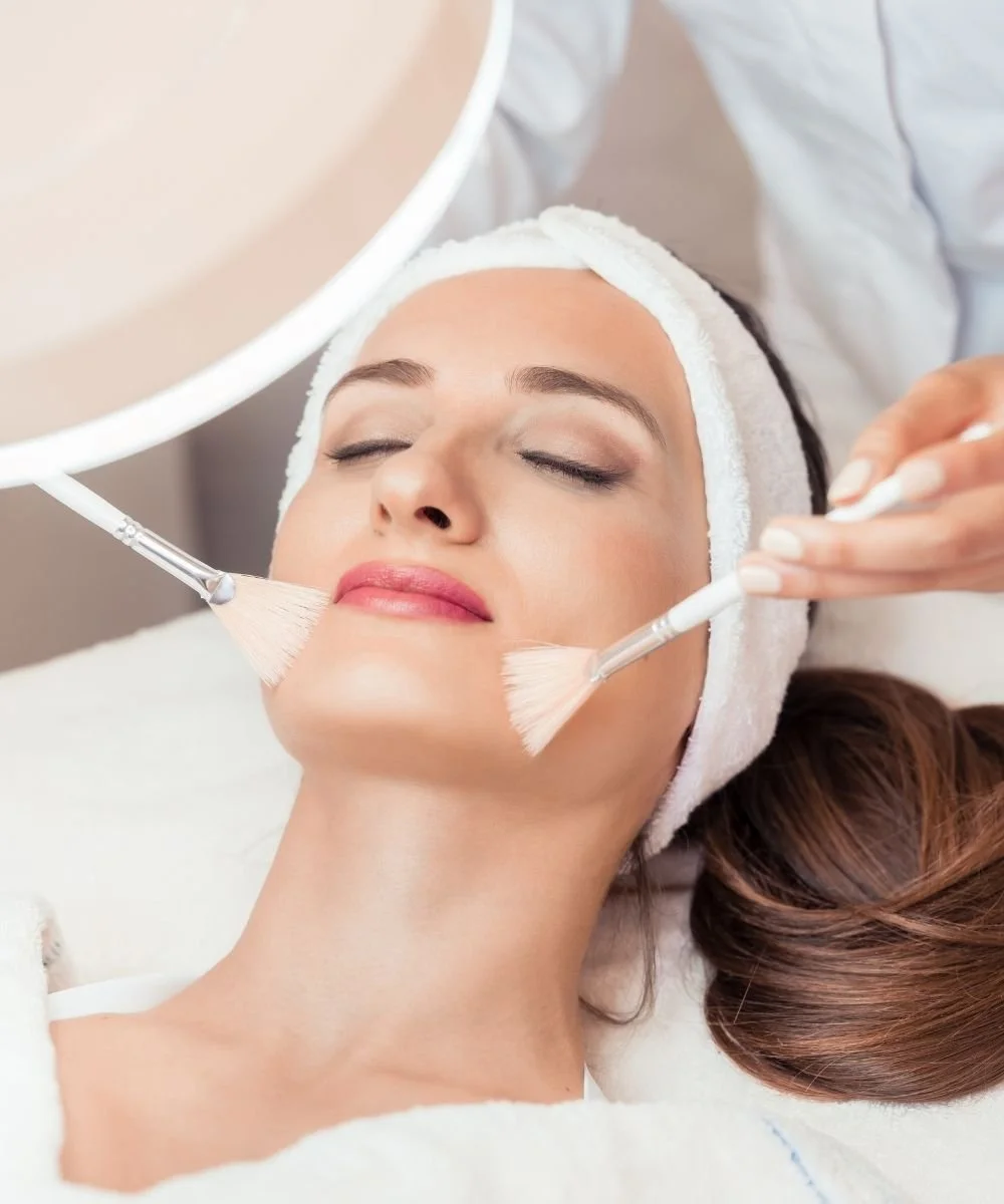 Close-up of woman receiving a facial treatment with brushes, lying down with a white headband, eyes closed, in a spa or skincare clinic.