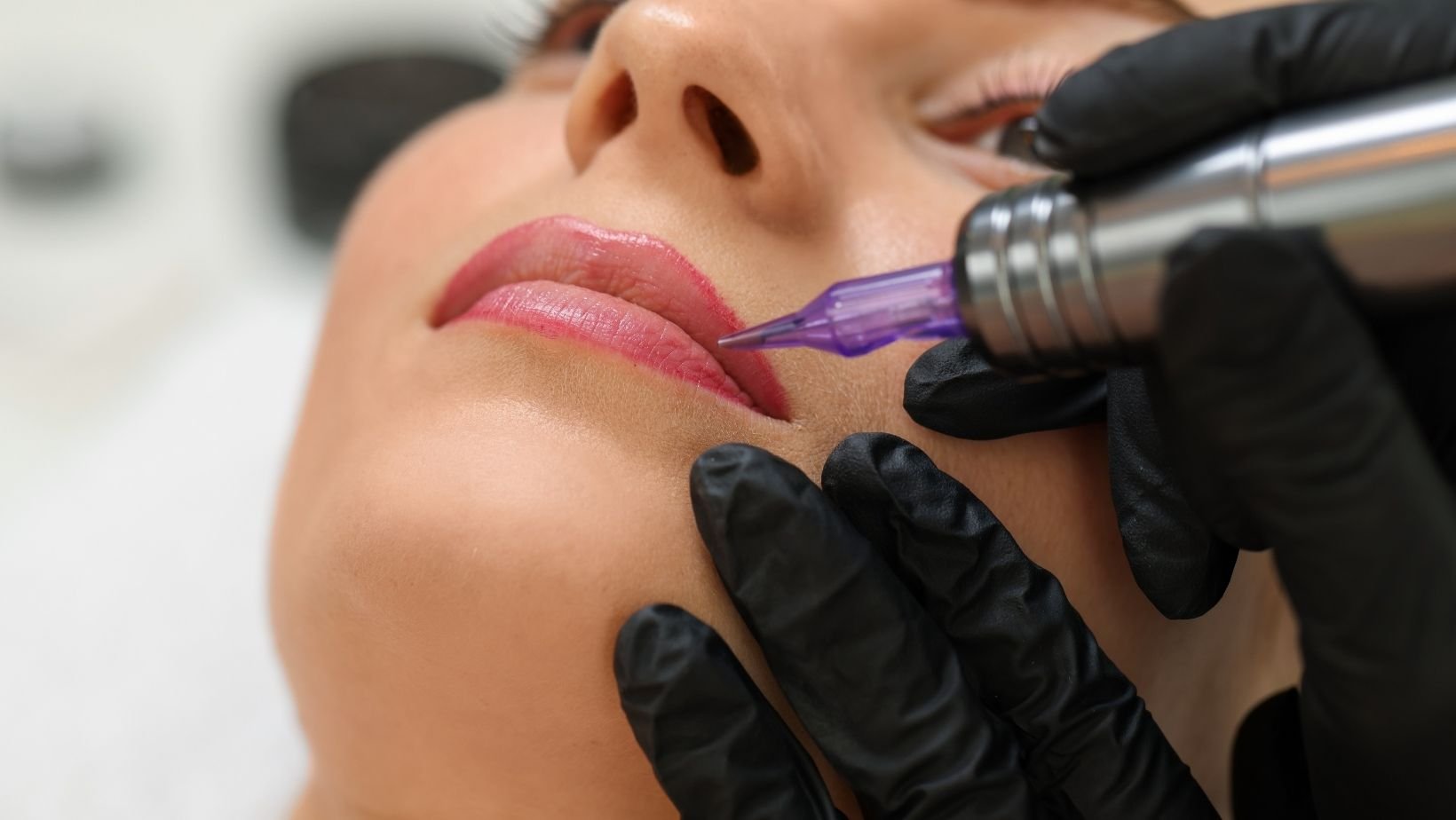 A person receiving a lip tattoo or lip blush procedure, with a professional using a tattoo machine on their lips.