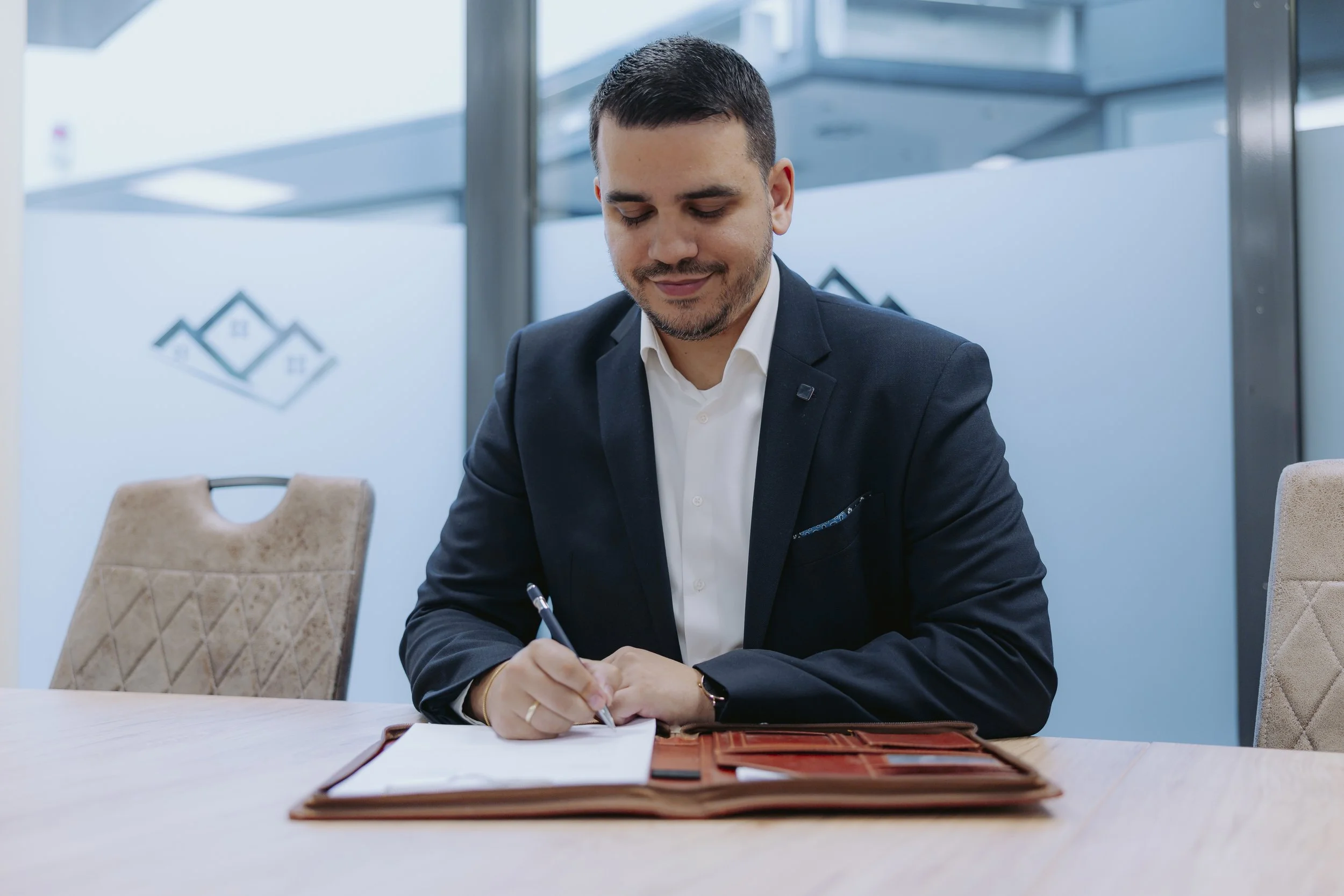 Der Mann sitzt an einem Tisch, schreibt in ein Notizbuch, trägt einen schwarzen Anzug und ein weißes Hemd in einem modernen Büro.