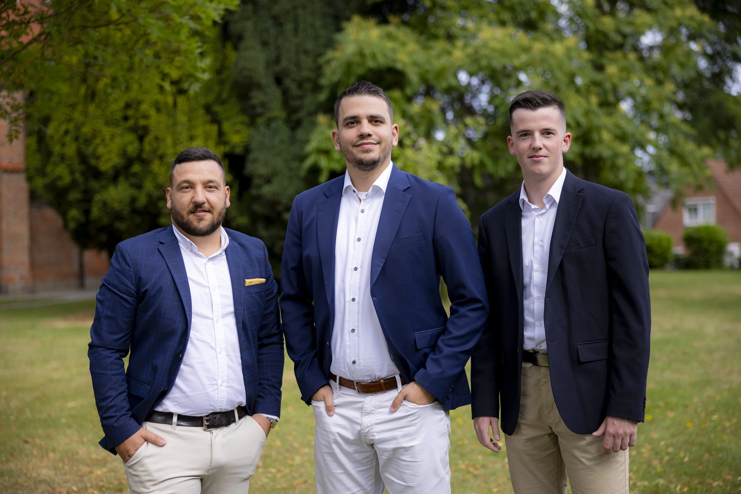 Drei junge Männer in Business-Kleidung stehen im Freien vor Bäumen und Häusern.