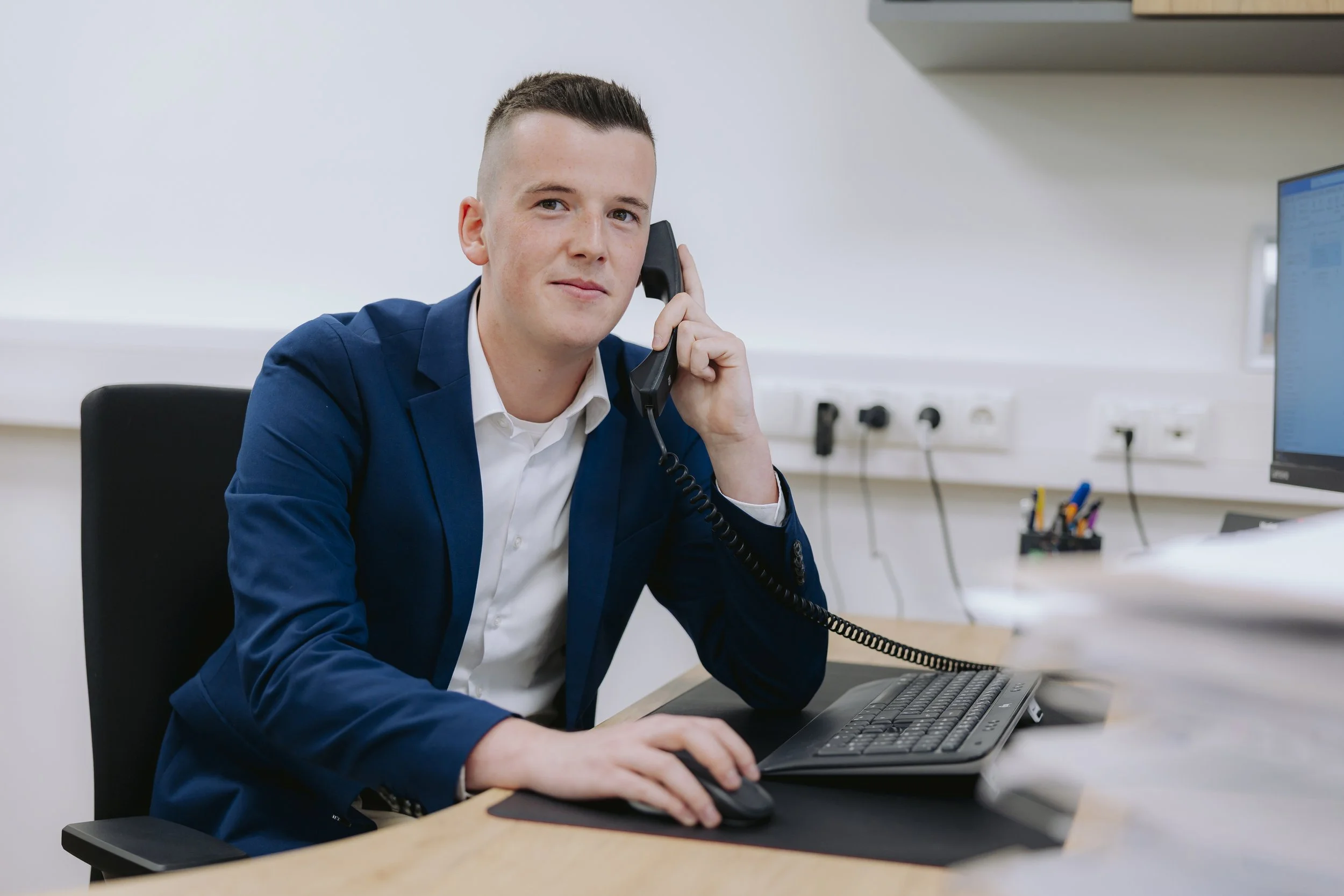 Ein junger Mann in Anzug sitzt an einem Schreibtisch, spricht am Telefon und nutzt die Computermaus.