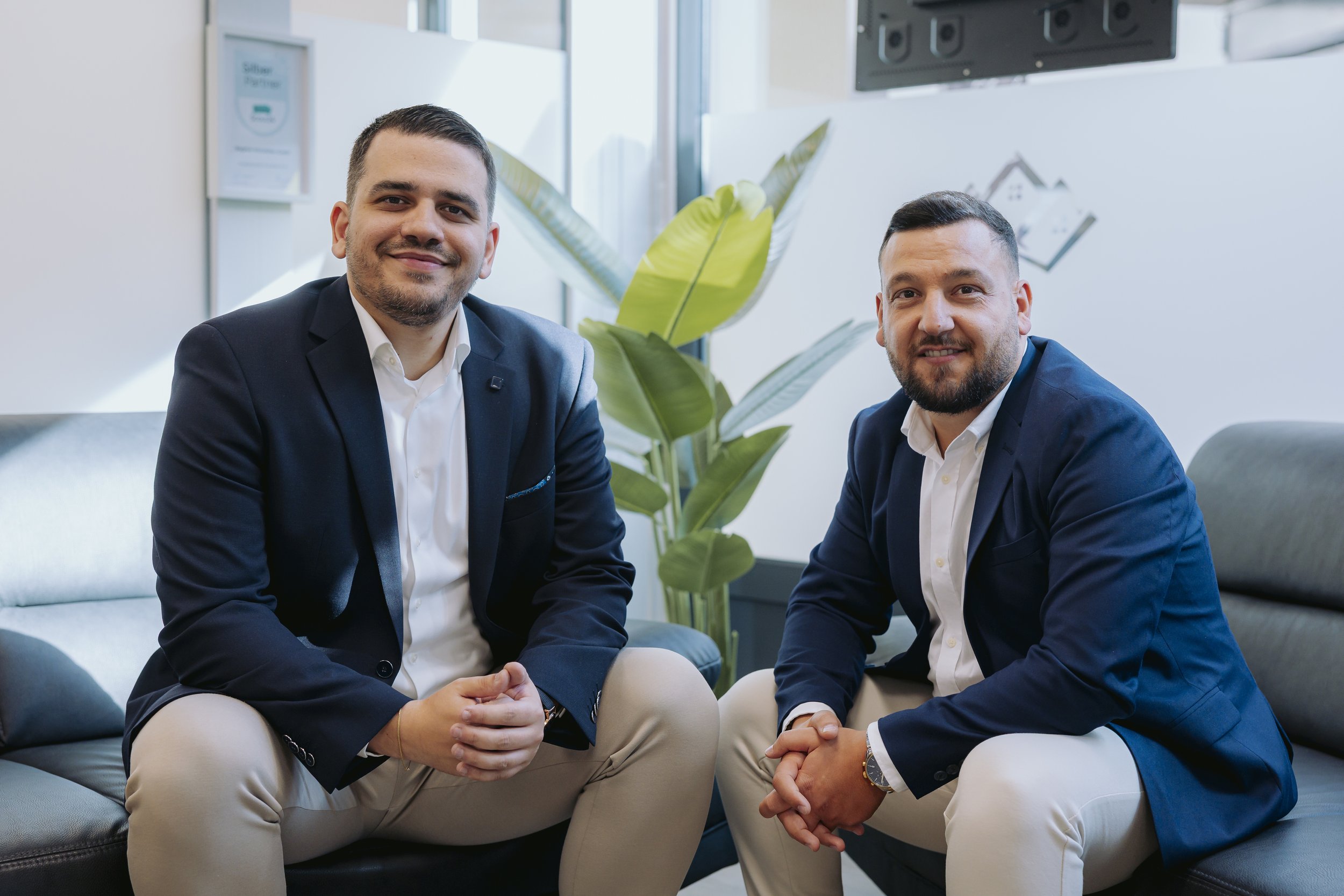 Zwei business gekleidete Männer sitzen auf einem Sofa in einem modernen Büro, lächeln und schauen in die Kamera, im Hintergrund eine Pflanze.