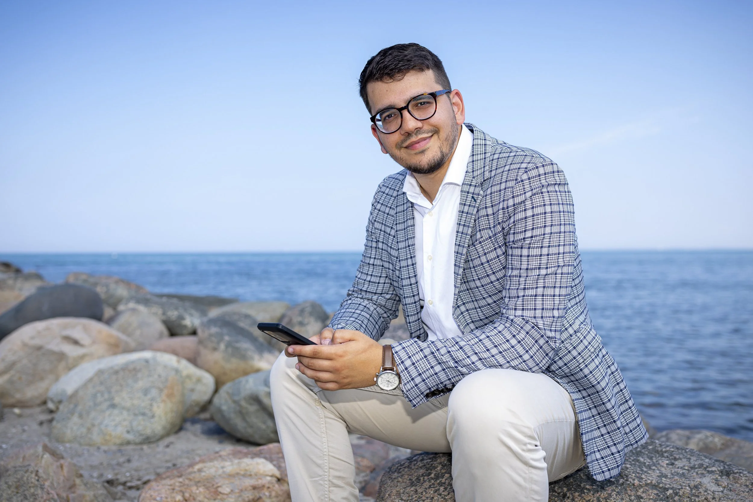 Ein Mann mit Brille sitzt an einem Ufer mit Steinen und hält ein Smartphone in der Hand, während er in die Kamera lächelt.