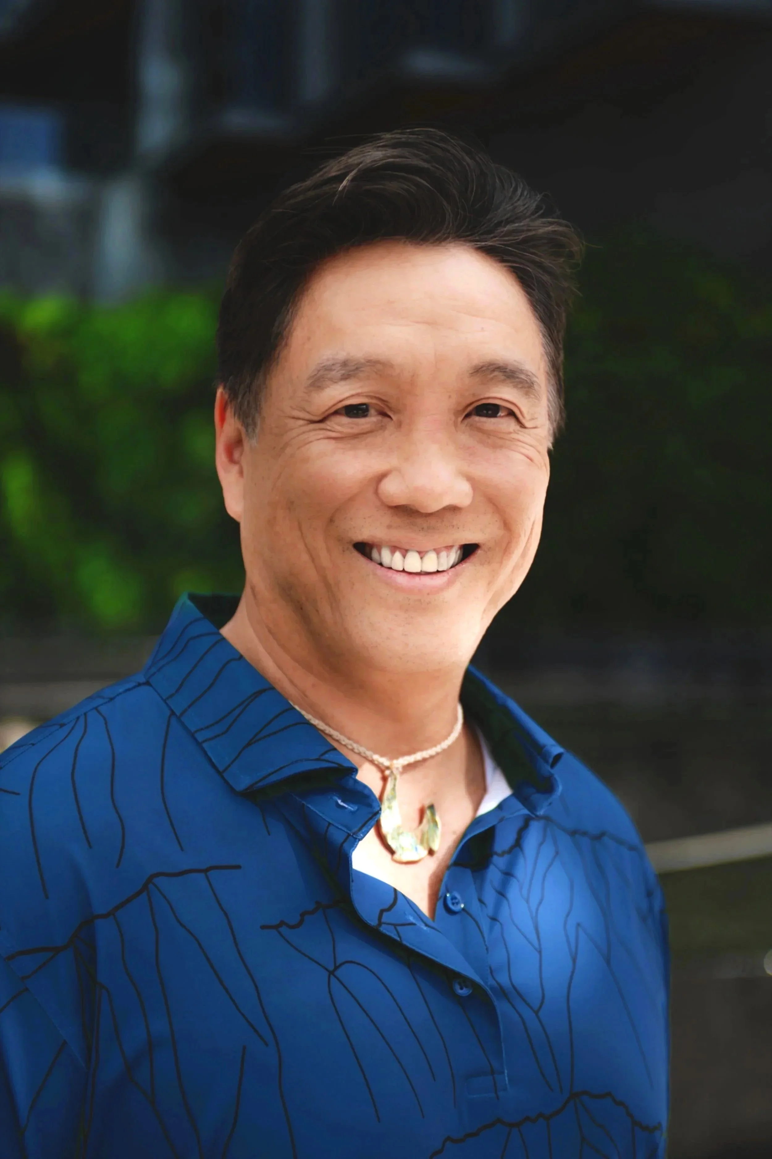 A smiling man wearing a blue shirt with thin black stripes, a white necklace, and a pendant, standing outdoors with a blurred background of greenery.