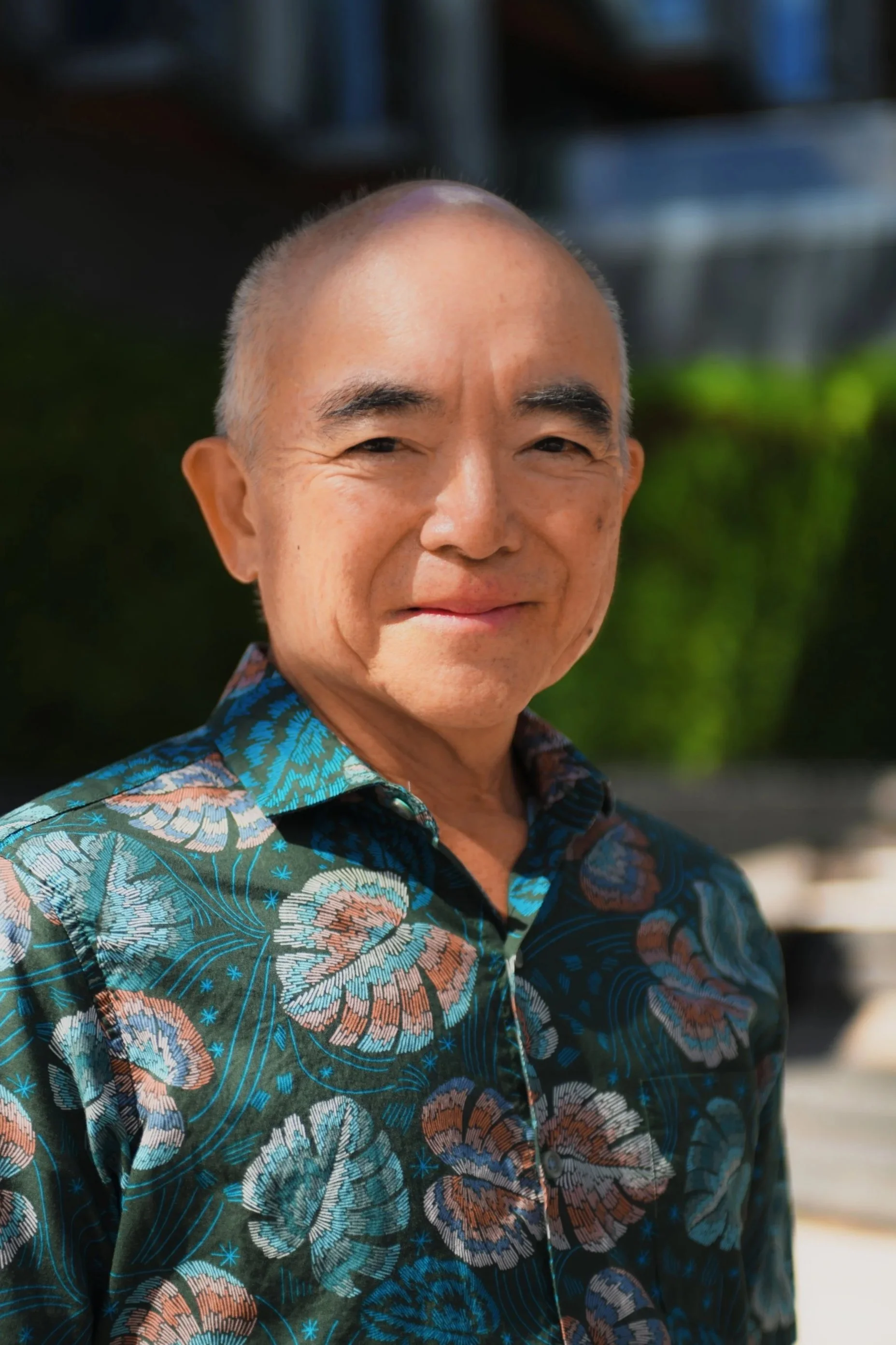 A man with a shaved head wearing a colorful Hawaiian shirt, smiling outdoors with a blurred background of greenery and buildings.