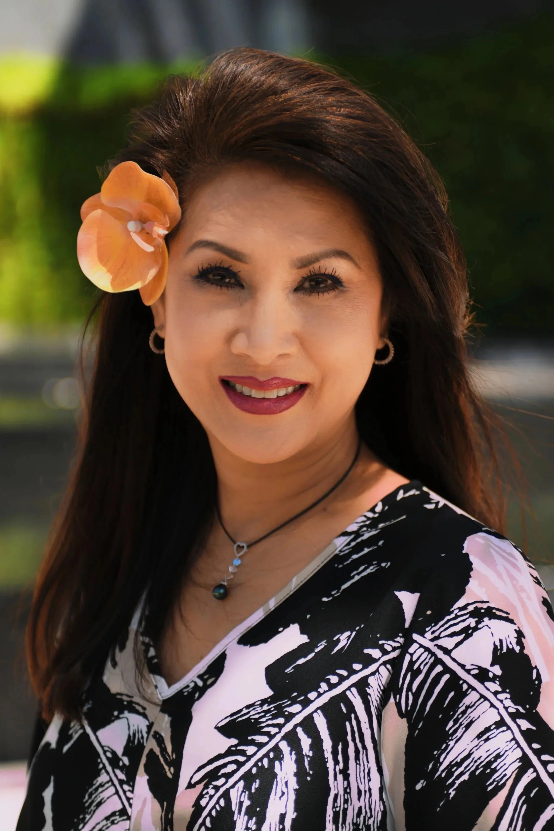 A woman with long dark hair and wearing makeup, a large peach-colored flower in her hair, earrings, a necklace, and a black and white patterned top, smiling outdoors with blurred greenery in the background.