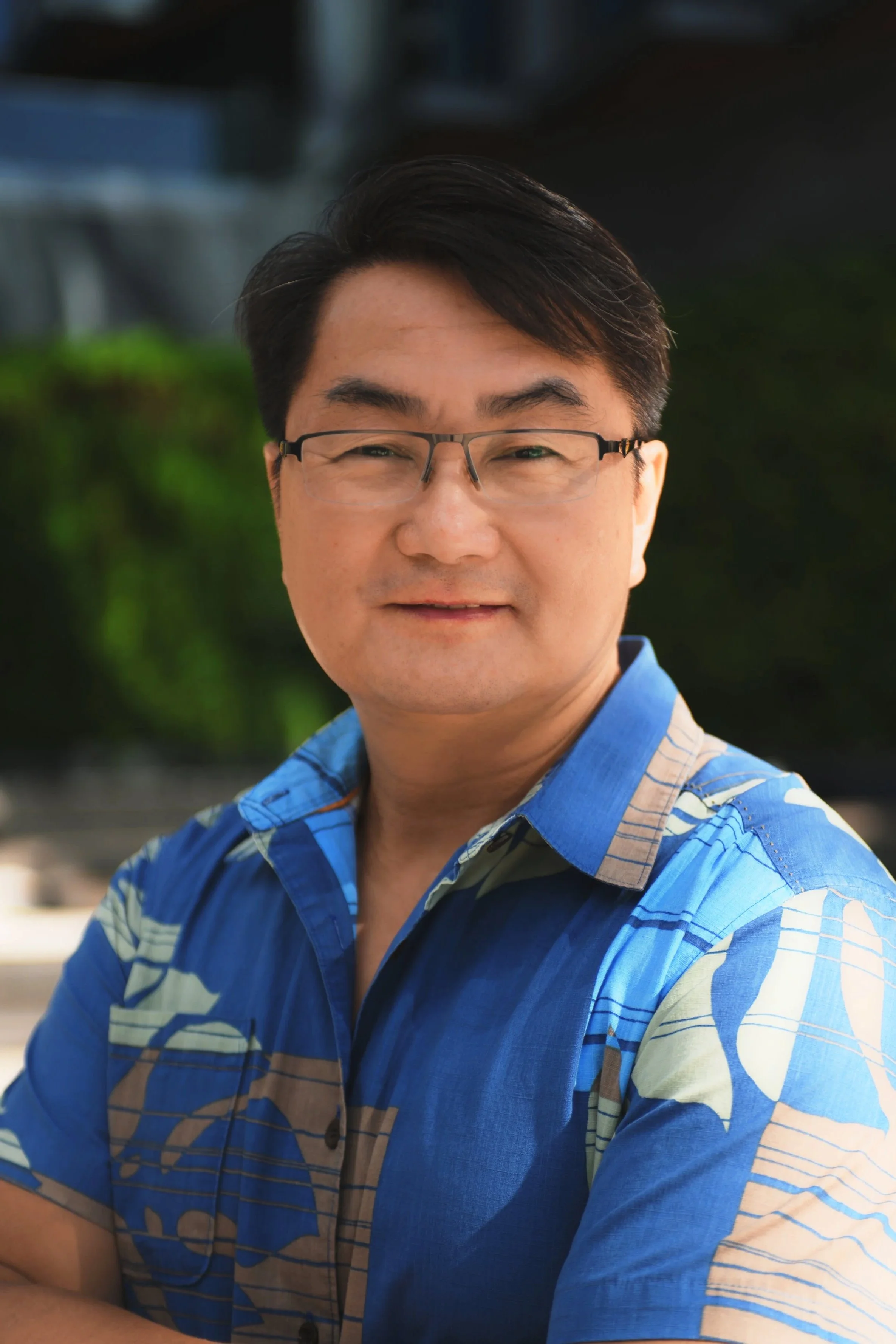 Close-up of a smiling man with glasses and short black hair, wearing a blue patterned button-up shirt, with blurred greenery background.