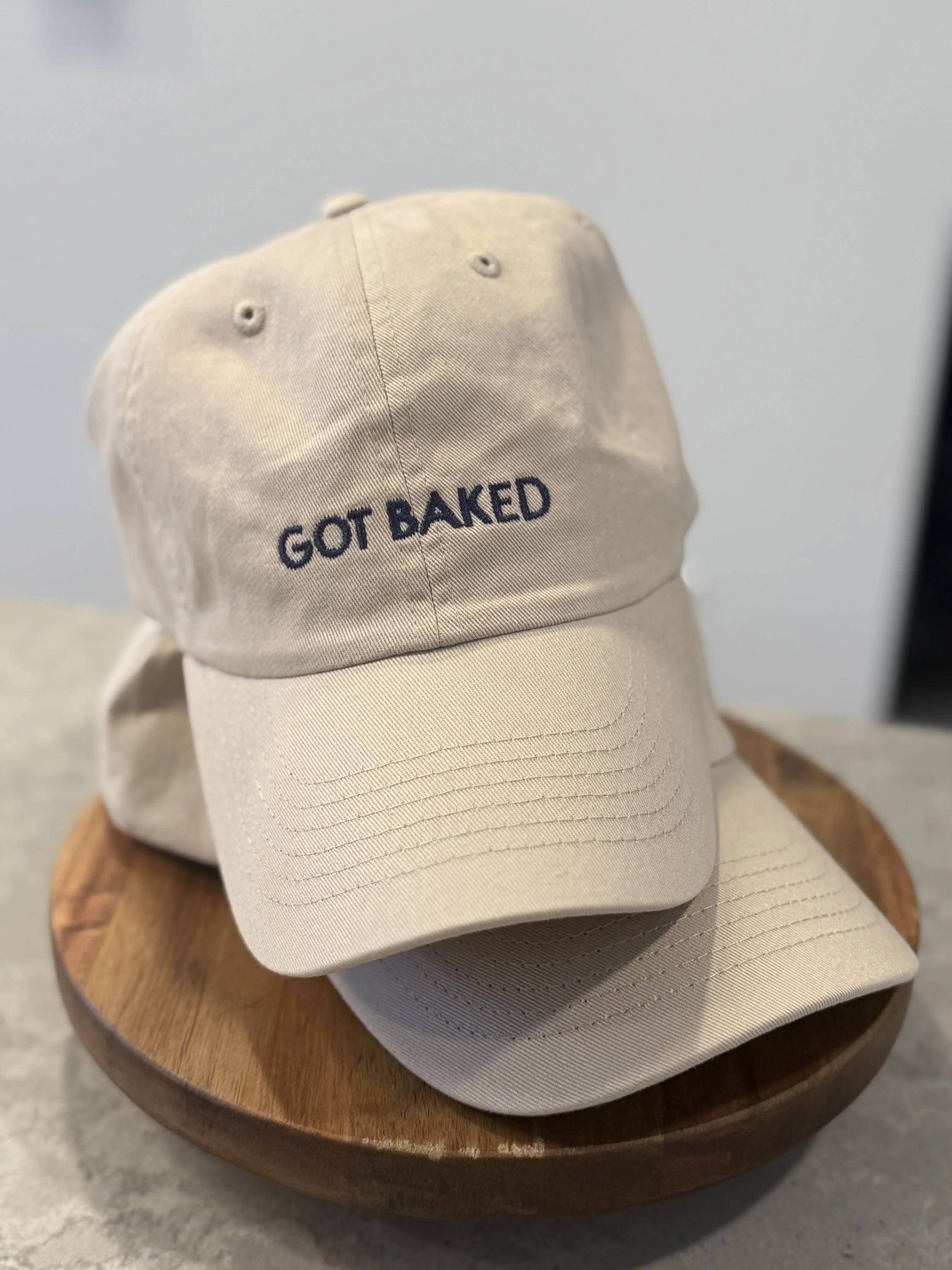 A beige baseball cap with the embroidered text 'GOT BAKED' on the front, resting on a round wooden surface.