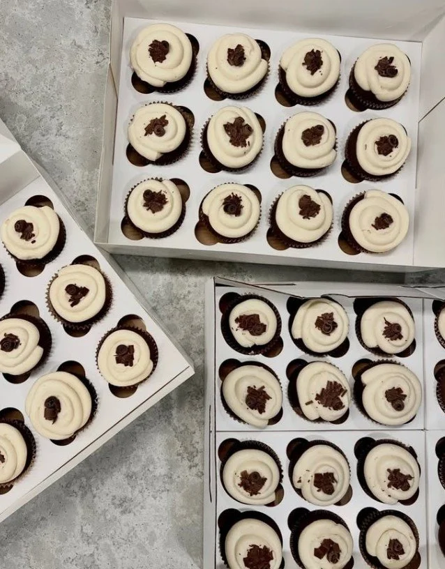 Boxes of chocolate cupcakes with white frosting and chocolate shavings on top.