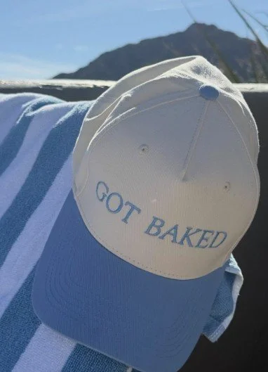 Beige baseball cap with blue embroidered text 'GOT BAKED' rested on a blue and white striped towel outdoors with mountains in the background.