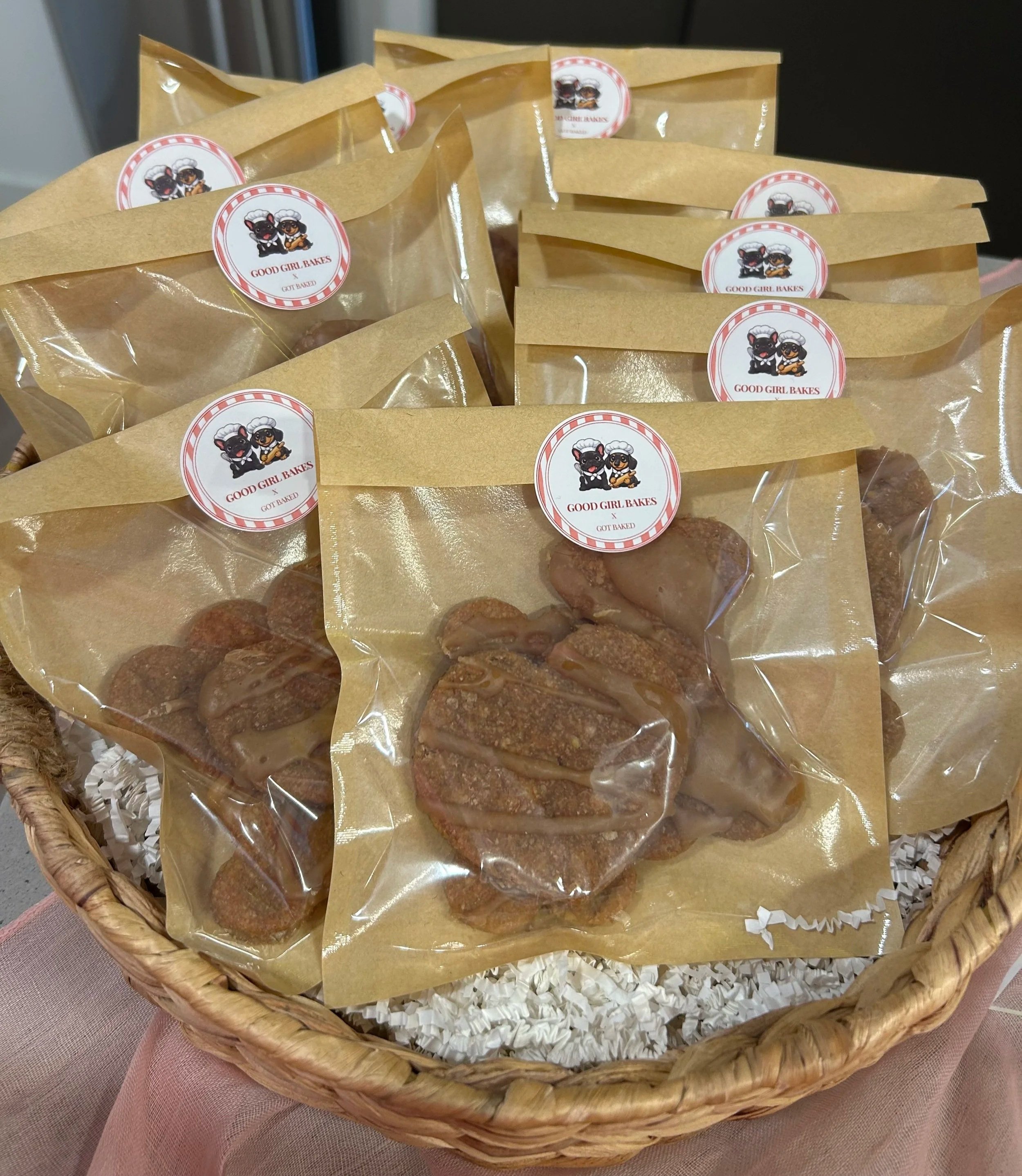 Assorted cookies in clear plastic bags with a red and white label reading "Good Girl Bakes" inside a wicker basket lined with white crinkle paper.