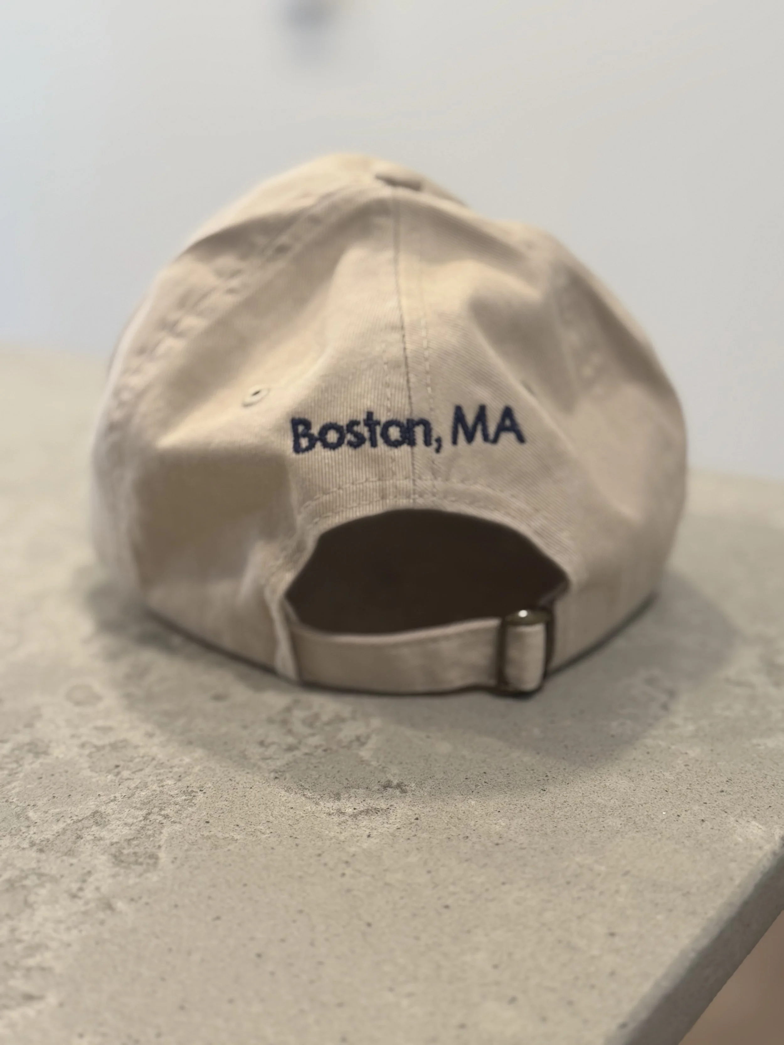 White baseball cap with navy embroidery reading 'Boston, MA' on the back, placed on a gray surface with a light background.