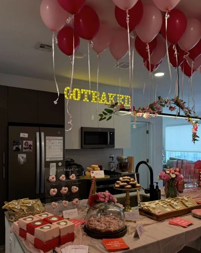 Birthday celebration setup with pink and red balloons hanging from the ceiling, a "COTBAKED" sign made of lights, a table with various baked goods, pink flower arrangements, and pink donut cookies displayed on a stand.