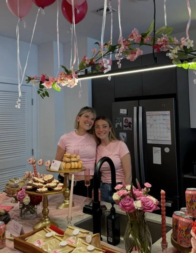 Two women smiling in a kitchen decorated with pink balloons, flowers, and birthday treats, celebrating a festive occasion.