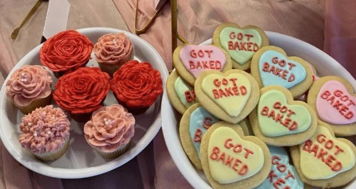 Cupcakes decorated with pink, red, and pinkish-beige frosting, alongside heart-shaped cookies with pastel-colored icing and 'GOT BAKED' written on them.
