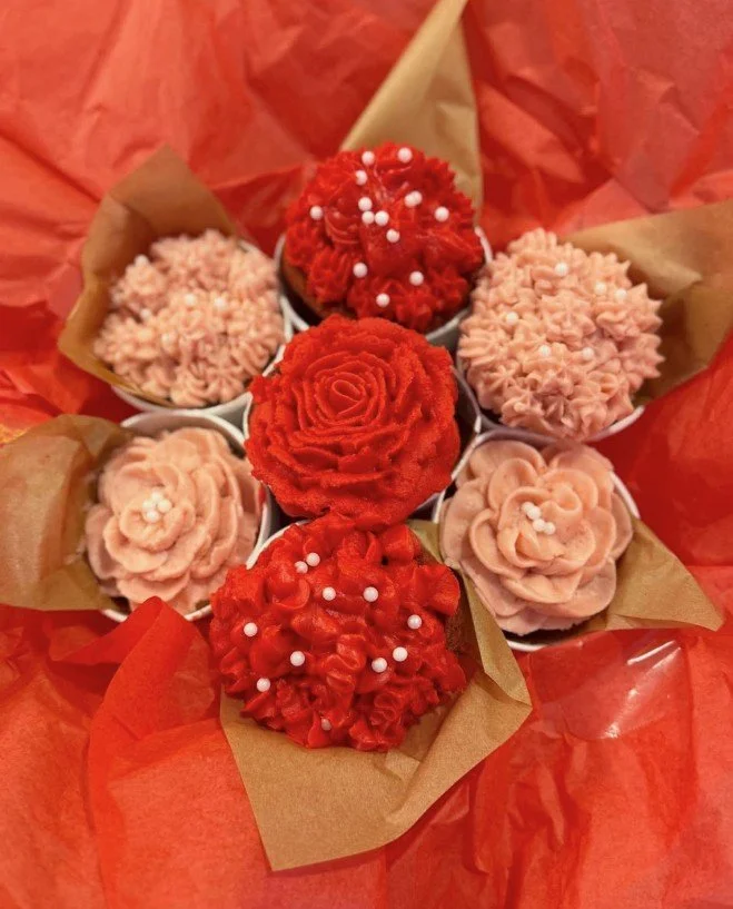 Cupcakes decorated with pink and red frosting, topped with small white sprinkles, arranged on tissue paper with a red wrapping background.