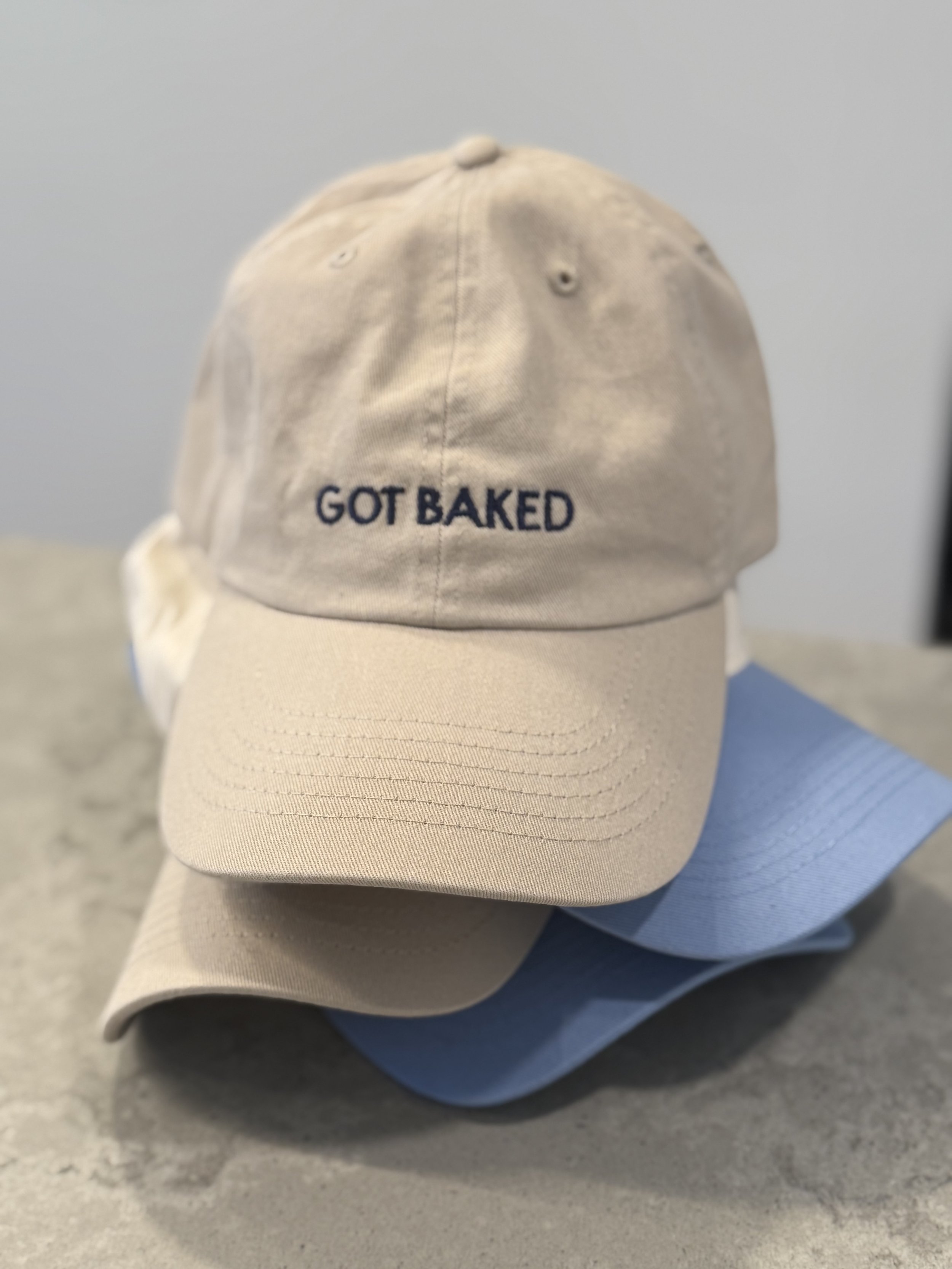 A beige baseball cap with the words "GOT BAKED" embroidered in navy blue on the front. The cap is resting on a person's lap, who is wearing light blue jeans and a white shirt.