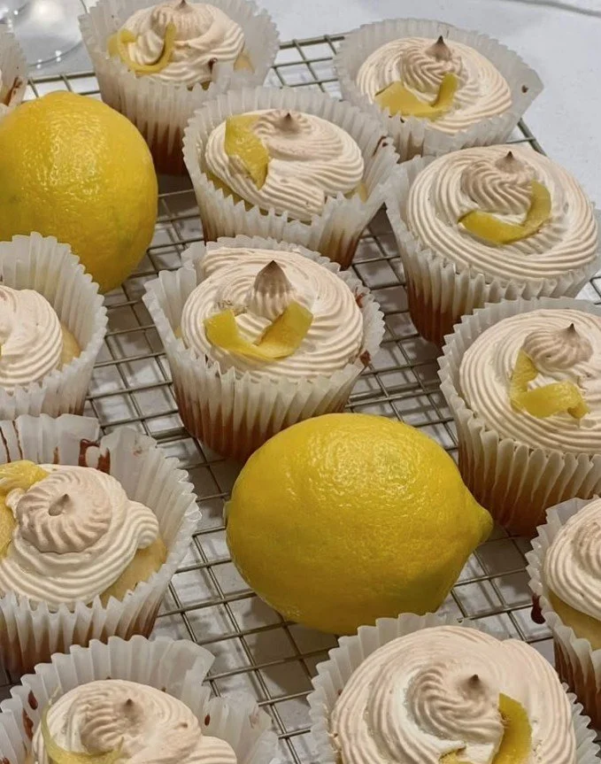 Lemon cupcakes with swirled white frosting and lemon zest garnishes, placed on a cooling rack with whole fresh lemons nearby.