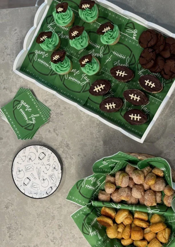 A tray of football-themed cupcakes with green frosting and chocolate top decorations, a plate of powdered sugar-covered fried snacks, theme-appropriate football napkins, and football-shaped cookies on a gray surface.