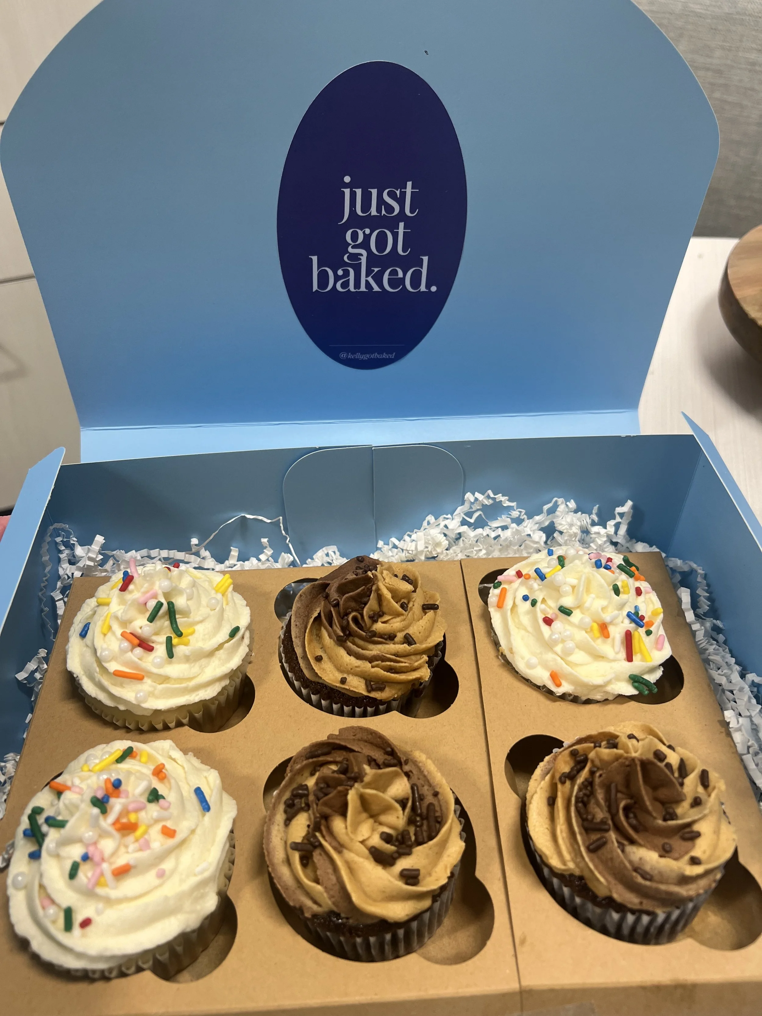 A box of six cupcakes with frosting and colorful sprinkles, chocolate and caramel toppings, in black paper liners, inside a blue bakery box.