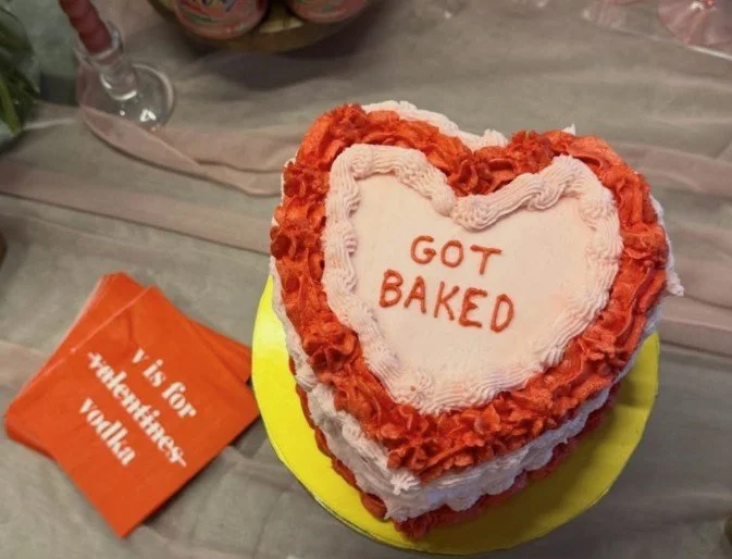Heart-shaped cake with red and pink frosting, decorated with the message 'GOT BAKED' written in red icing, sitting on a yellow plate. There is an orange card on the table next to the cake that reads 'V is for Valentine, Valentine'.