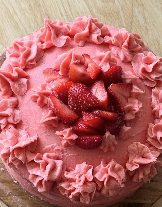 A round pink strawberry cake decorated with pink whipped cream ruffles around the edge and topped with sliced fresh strawberries in the center.