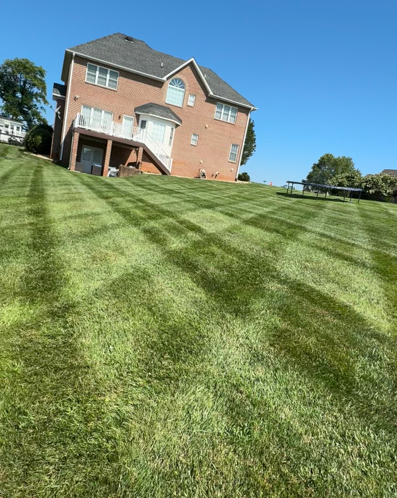 Backyard with freshly mowed green grass, a brick house with a deck, and a trampoline on the lawn under a clear blue sky.