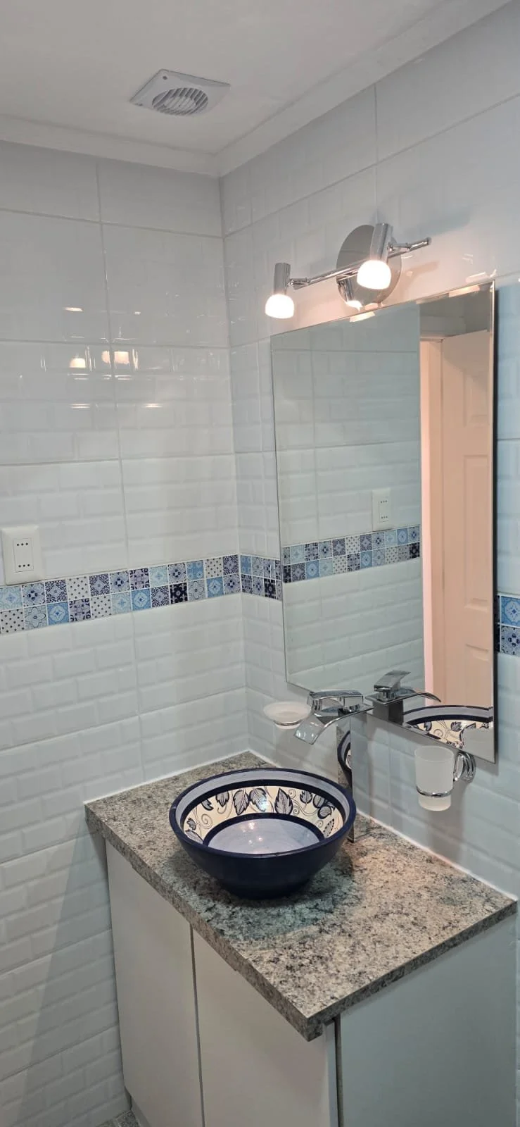 Lavabo de cerámica decorado con diseño floral, granito en la encimera, espejo grande y luz de pared en un baño con azulejos blancos y detalle decorativo azul.