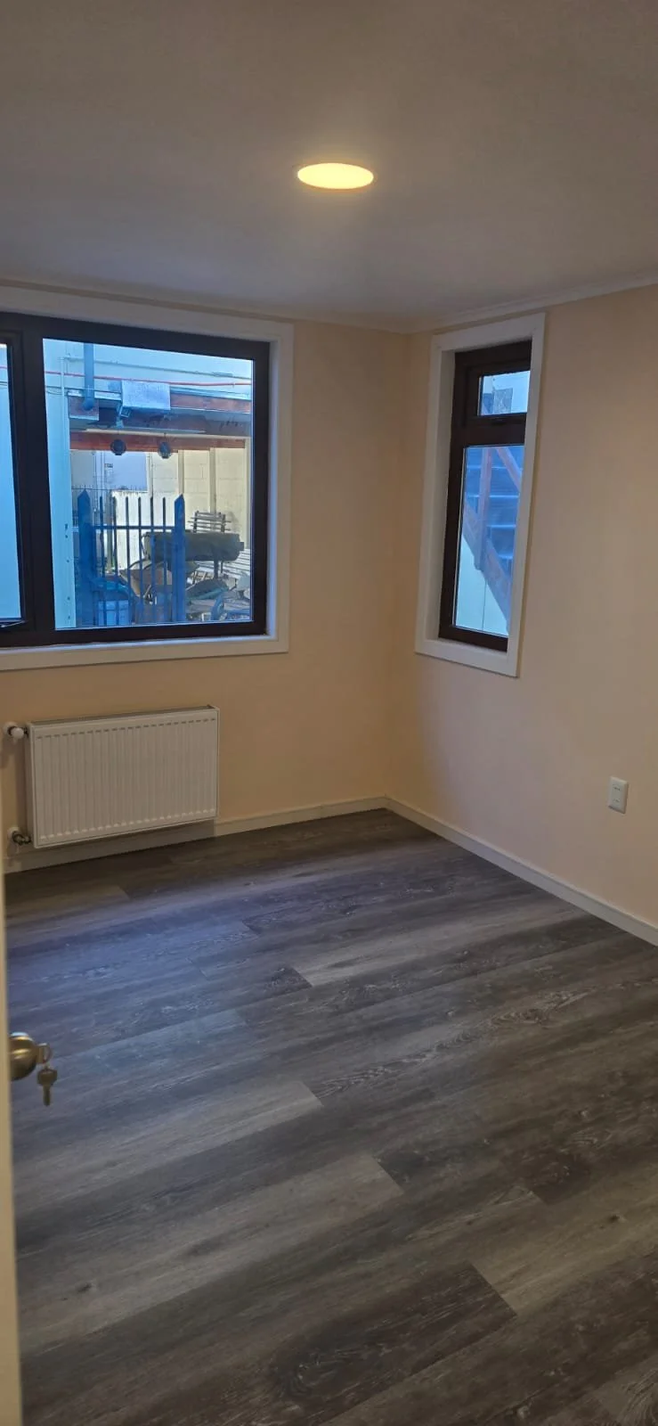 Habitación vacía con paredes color beige, dos ventanas con vista al exterior, suelo de laminado en tonos grises, radiador en la pared y luz de techo encendida.