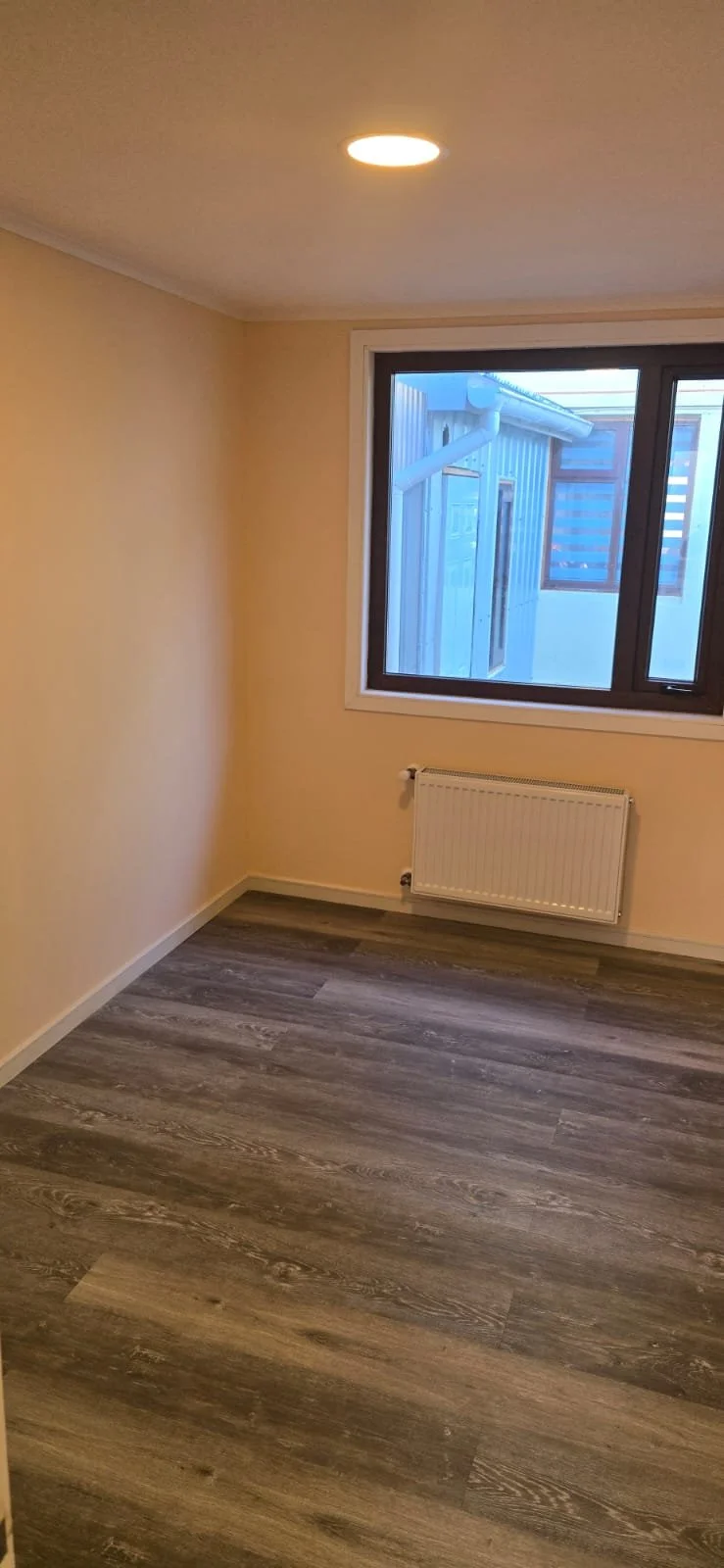 Habitación vacía con paredes crema, ventana grande, radiador y pisos de madera en tonos grises y marrones.