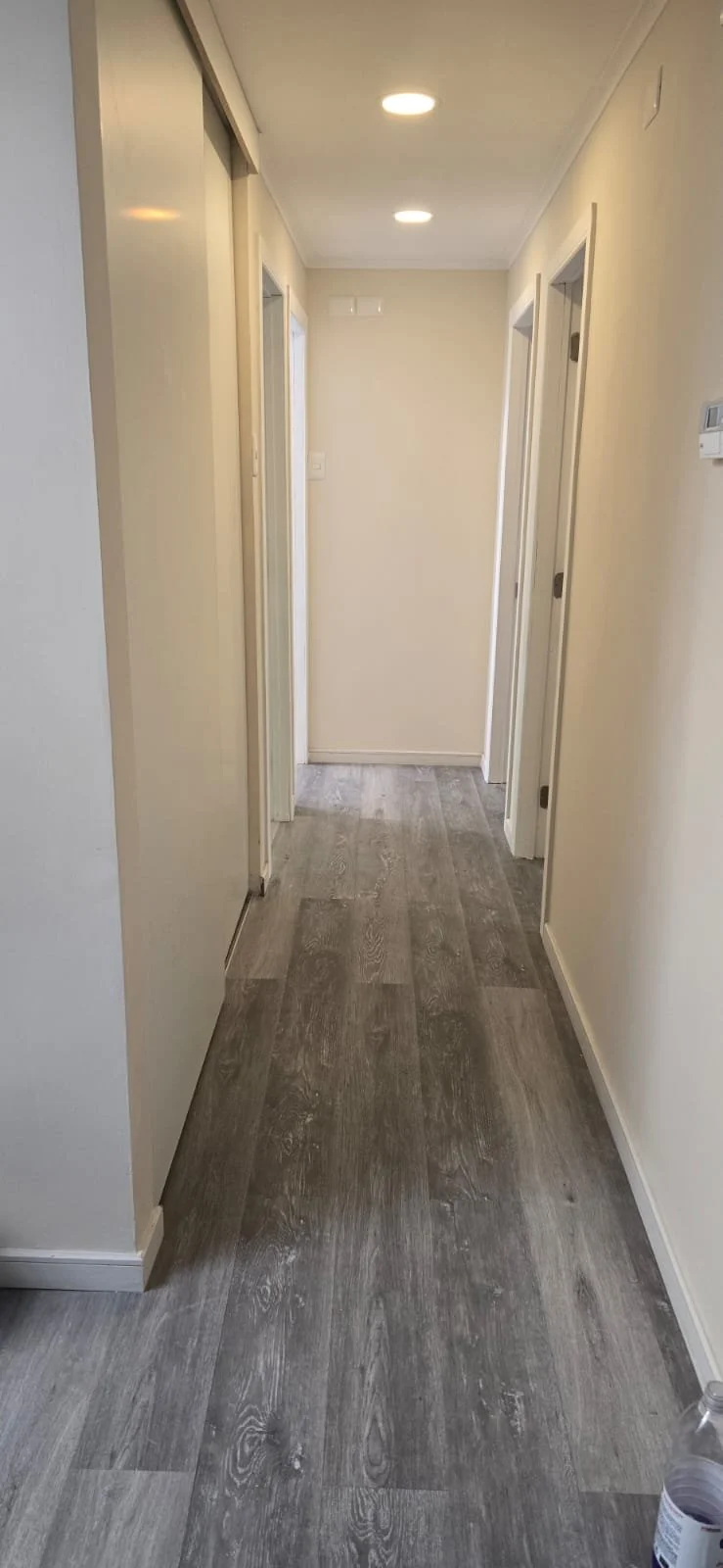 Pasillo interior con paredes blancas, puertas blancas, suelo de madera gris y luces empotradas en el techo.