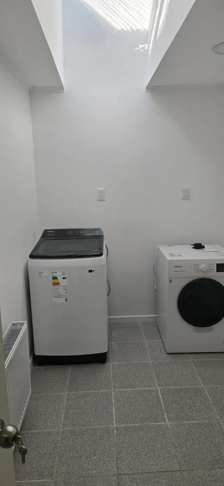 Lavandería con lavadora y secadora blancas en una habitación con piso de baldosas grises y paredes blancas, con un tragaluz en el techo.