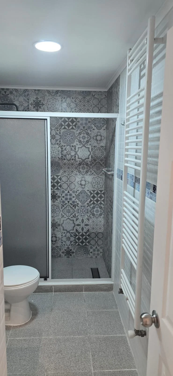 Vista interior de baño con váter, ducha con paredes de azulejos decorativos en tonos grises, ducha con puertas corredizas, y toallero blanco. Piso de baldosas grises.