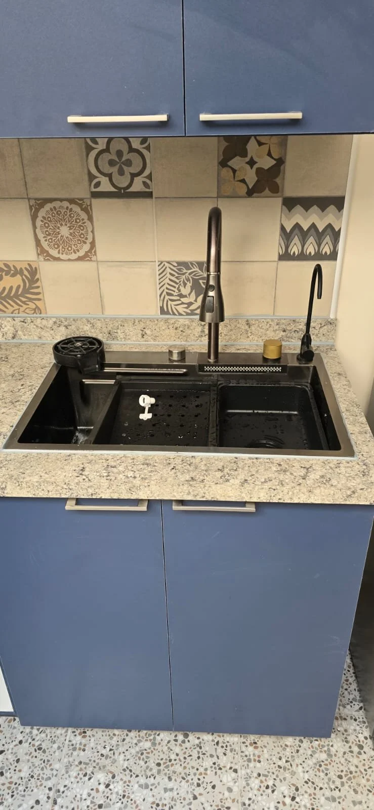 Fregadero de cocina con grifo, tapa de recipiente y electrodoméstico en tonos oscuros, sobre encimera de granito, con azulejos decorativos en la pared y gabinetes azules.