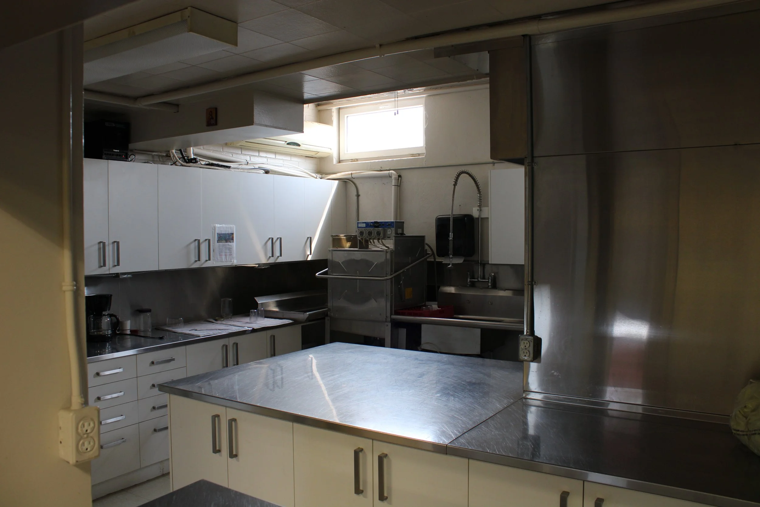 A compact commercial kitchen with white cabinets, stainless steel countertops, a dishwashing sink, and a small window letting in natural light.