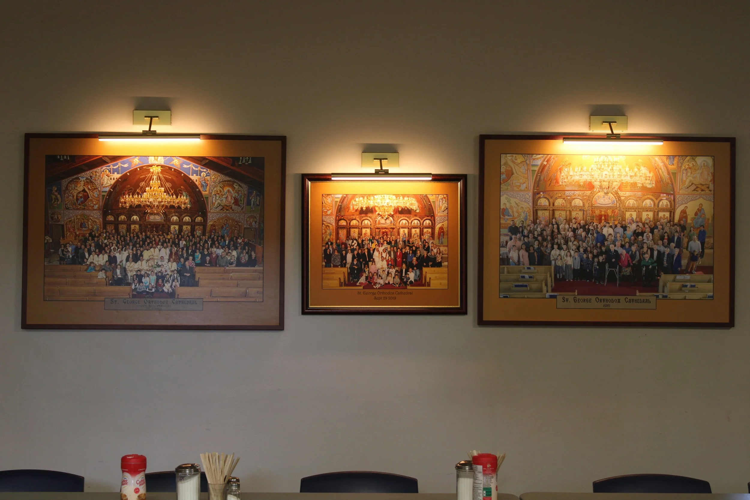 Three framed paintings of church interiors and congregations on a wall illuminated by three light fixtures, with a table below holding condiments, napkins, and utensils.