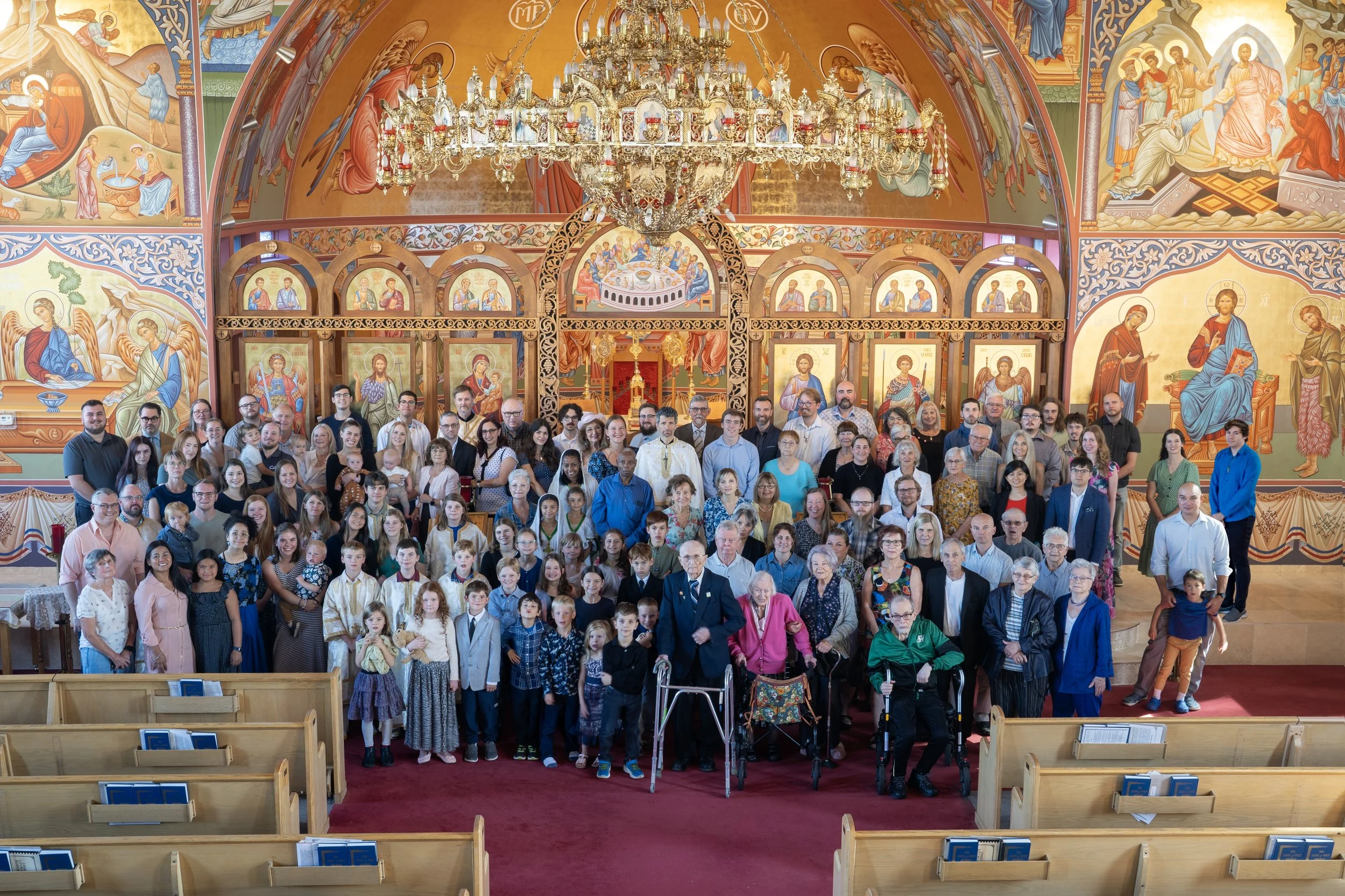 Large group of people gathered inside an ornate church decorated with religious icons and murals, gathered for a service or event.