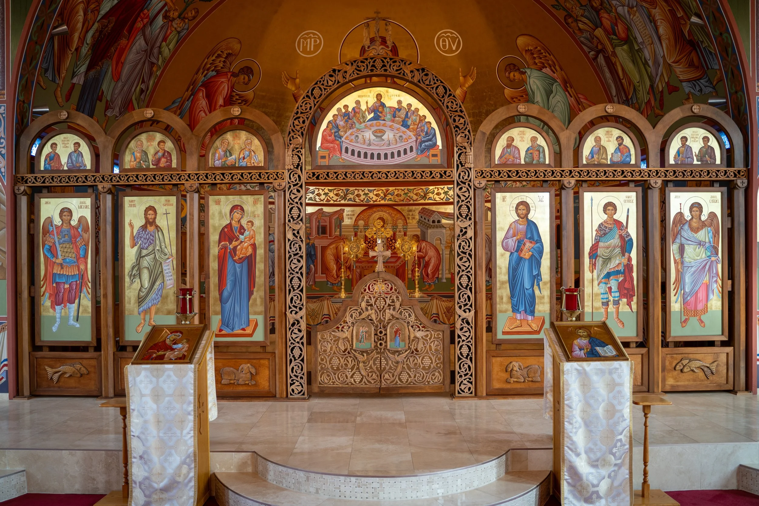 Inside a Christian church, an ornate altar with icons of saints and biblical scenes, including Jesus and the Virgin Mary, surrounded by intricate wooden carvings and religious imagery, on a gold and colorful backdrop.