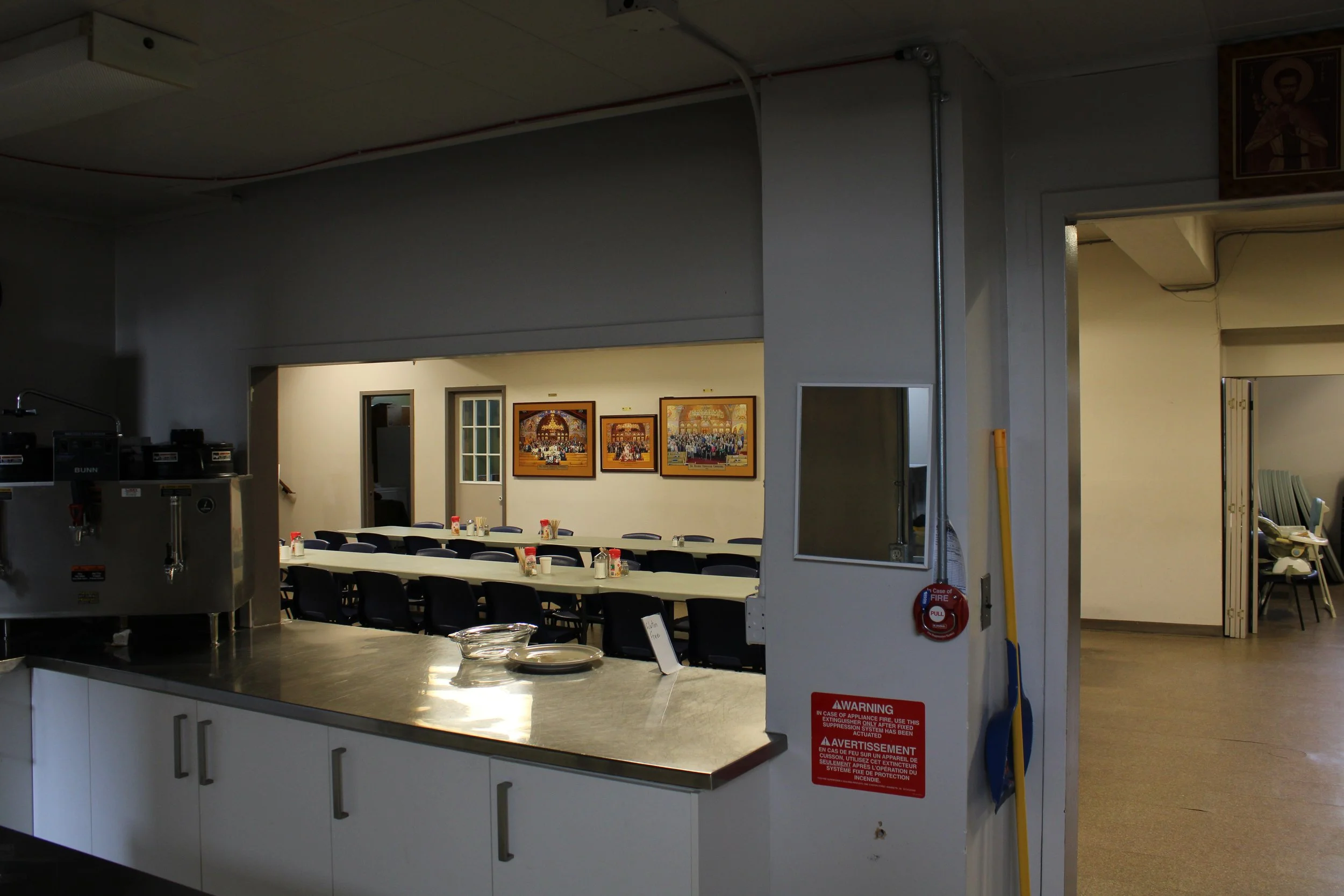 A cafeteria or dining area with a long table set with small condiment bottles and cups, behind a window opening in a wall, with framed pictures hanging on the wall in a well-lit room.