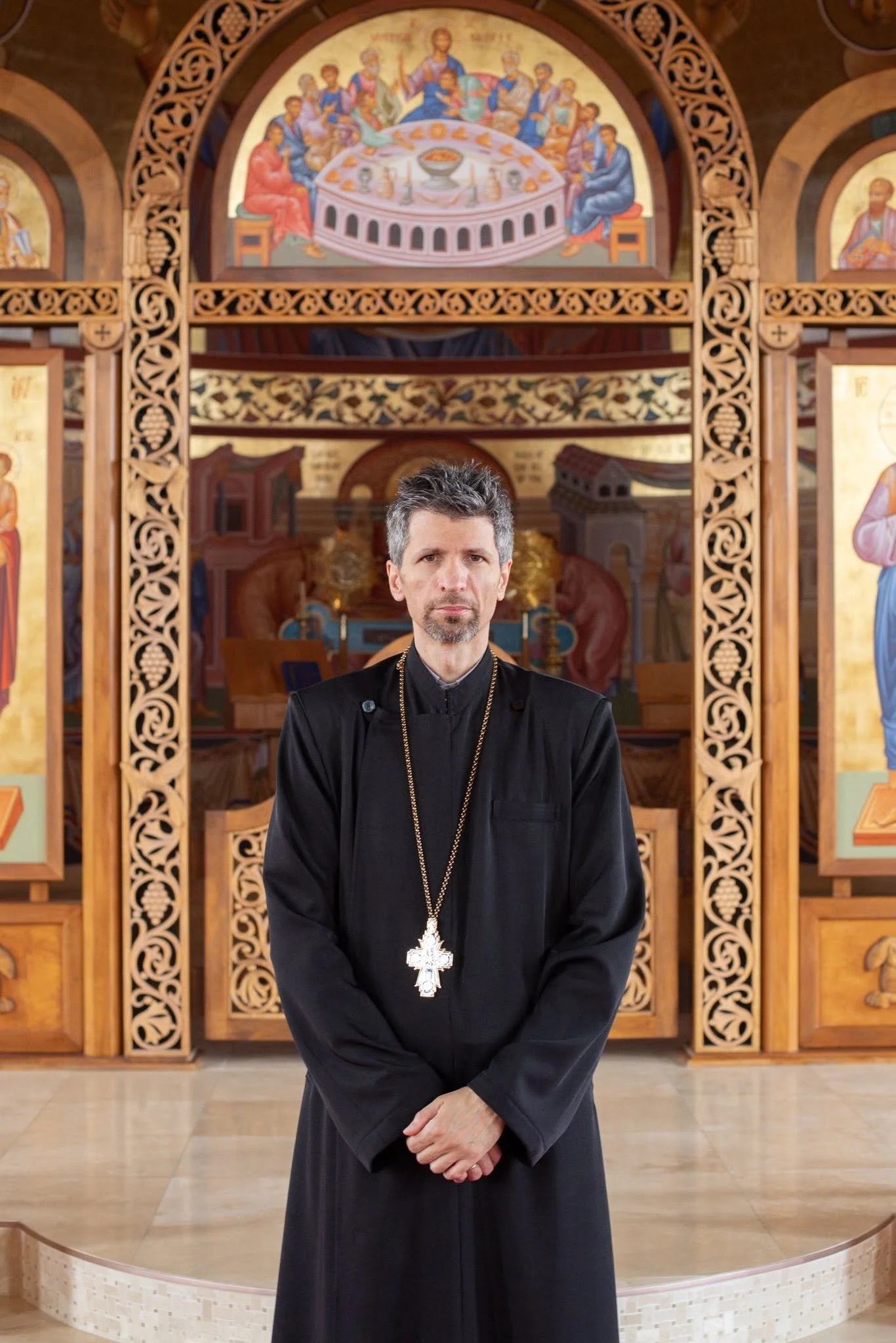 A man dressed in black religious attire with a large cross around his neck is standing in an ornate church with religious paintings on the wall behind him.