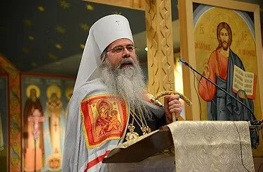 The Most Blessed Tikhon (Mollard) Archbishop of Washington Metropolitan of All America and Canada speaking at a religious service, wearing a white head covering and robes with religious icons, standing in front of icons of Jesus and saints.