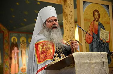 The Most Blessed Tikhon (Mollard) Archbishop of Washington Metropolitan of All America and Canada speaking at a religious service, wearing a white head covering and robes with religious icons, standing in front of icons of Jesus and saints.