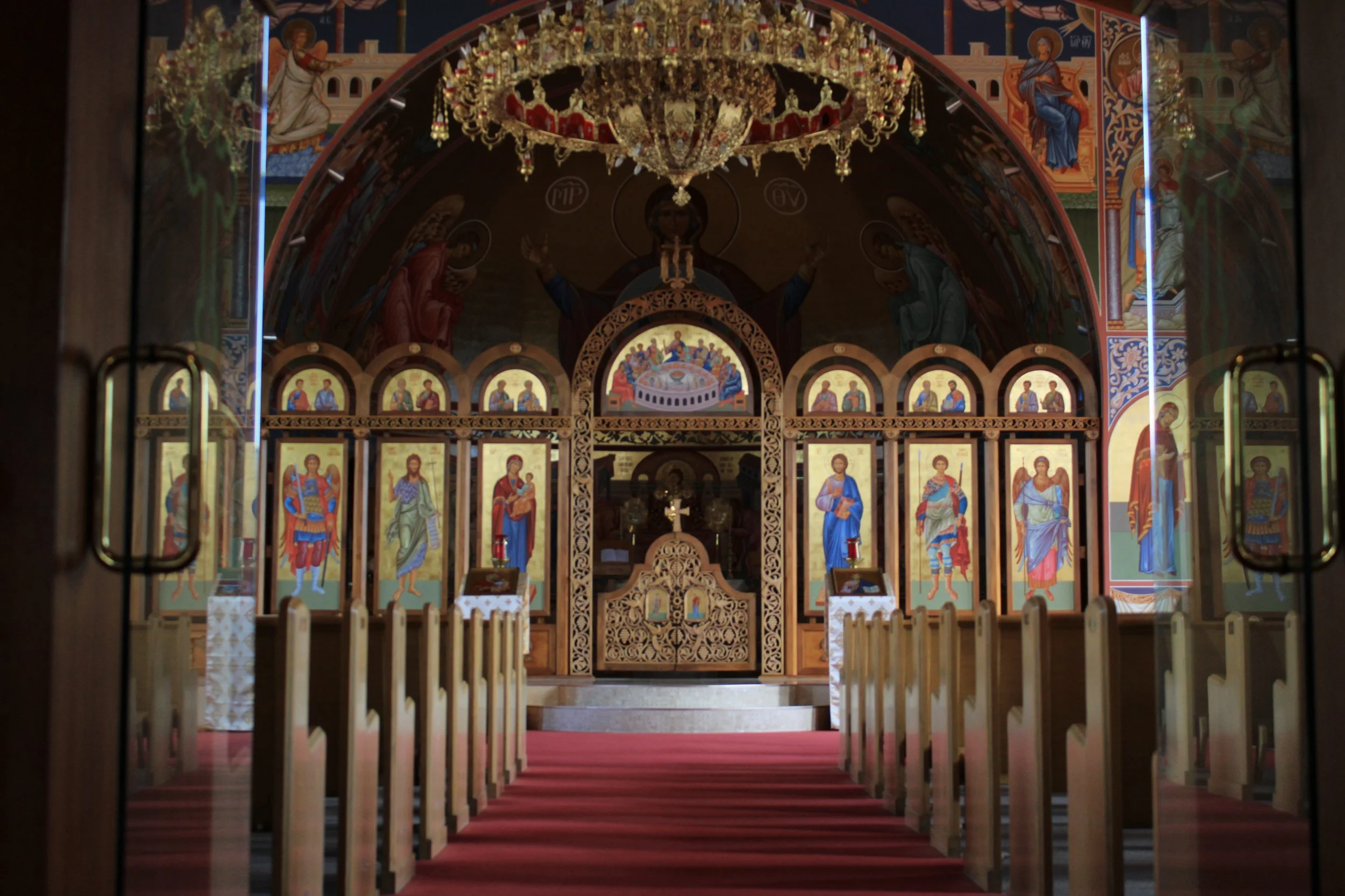 The interior of an Orthodox Christian church with religious icons, ornate decorations, and a chandelier hanging from the ceiling. Wooden prayer stands are front and center, and the view is through a doorway with metal handles.