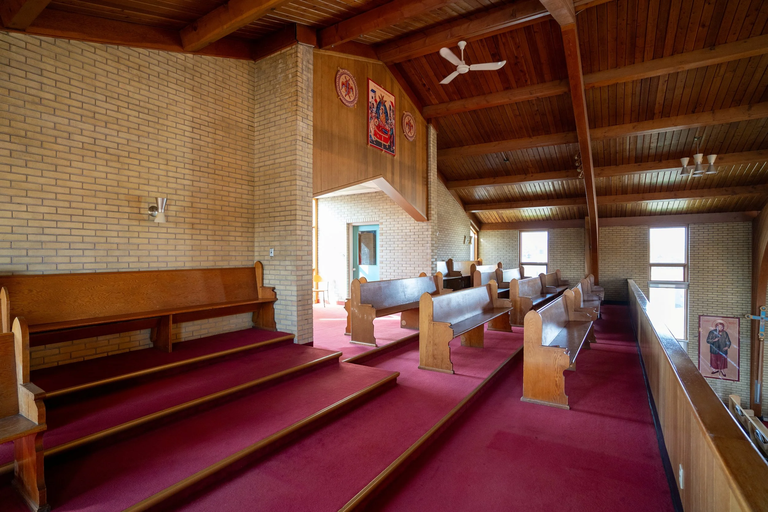 Interior view of a church with wooden wall paneling and a vaulted wooden ceiling, red carpeted steps, and pews. Religious icons and banners are displayed on the walls, and windows let in natural light.