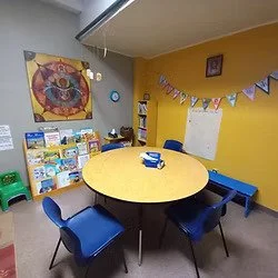 Saint George Orthodox Cathedral youth group activity room with a yellow wall and a round table surrounded by six blue chairs. The wall features bookshelves, a colorful art piece, and a patriotic banner. There are some toys and books on the shelves.