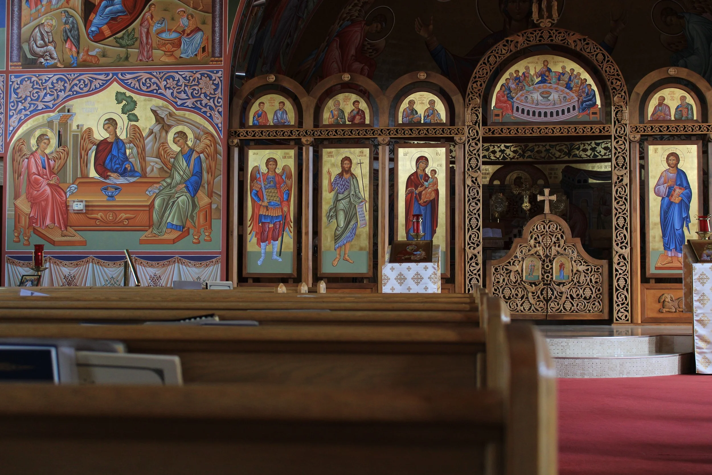 Inside a church with religious icons and paintings on the wall behind a wooden altar, featuring depictions of saints and biblical scenes, with a red carpeted floor.
